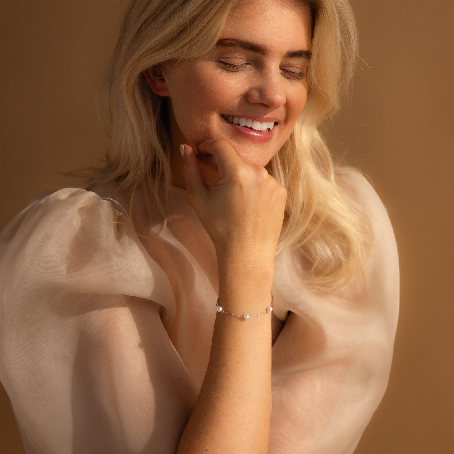 A smiling woman with long blonde hair, wearing a sheer puff-sleeve blouse and accessorized with the modern Pearl Station Bracelet.