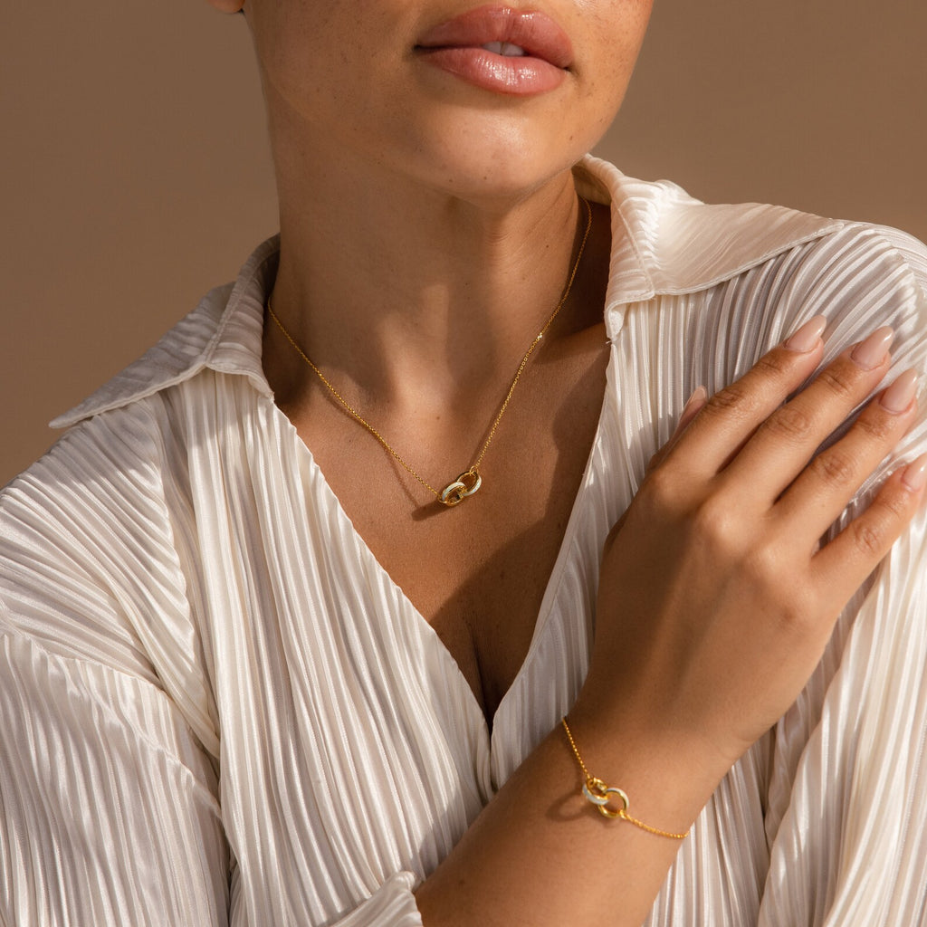 Woman in a white pleated blouse wears the Interlocking Opal Inlay Necklace with matching bracelet, hand resting on chest—a symbol of infinite love and an ideal anniversary gift.