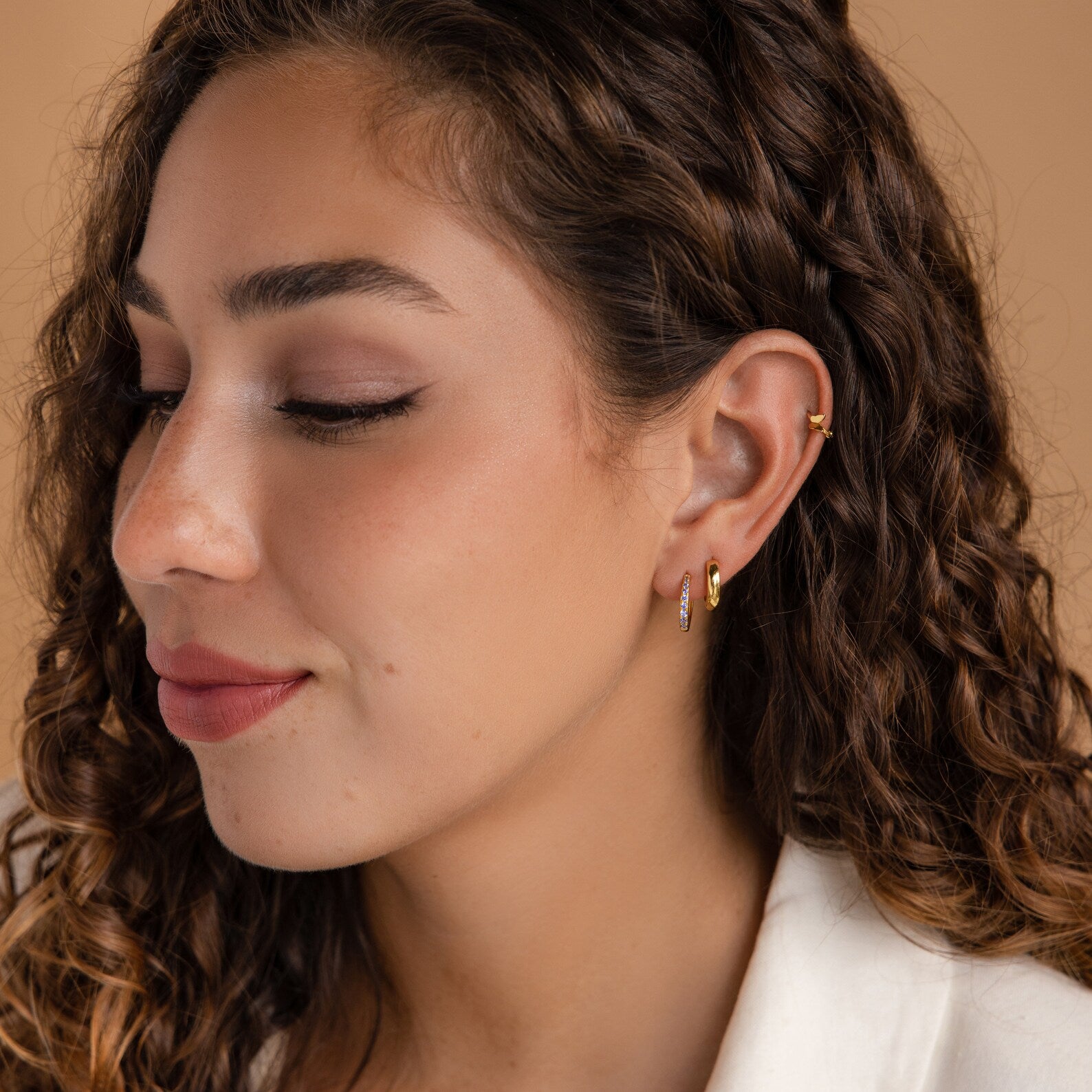 Woman with curly brown hair and gold hoop earrings, wearing a white top, faces left against a beige background. Her Vera Ruby Birthstone Huggies add a subtle pop of color to her classic look.