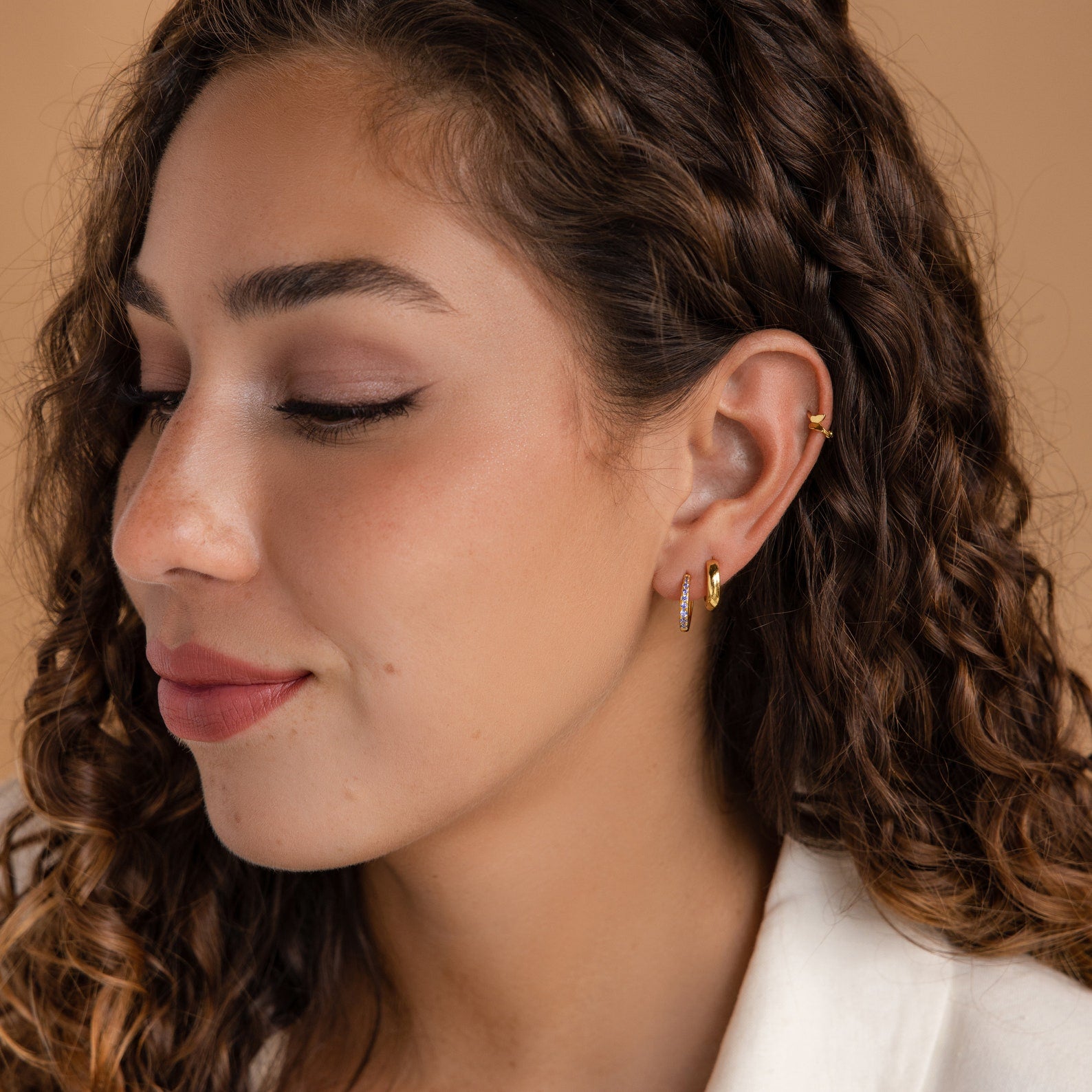 Woman with curly brown hair wearing the Vera Emerald Birthstone Huggies and a white top, shown in a soft side profile.