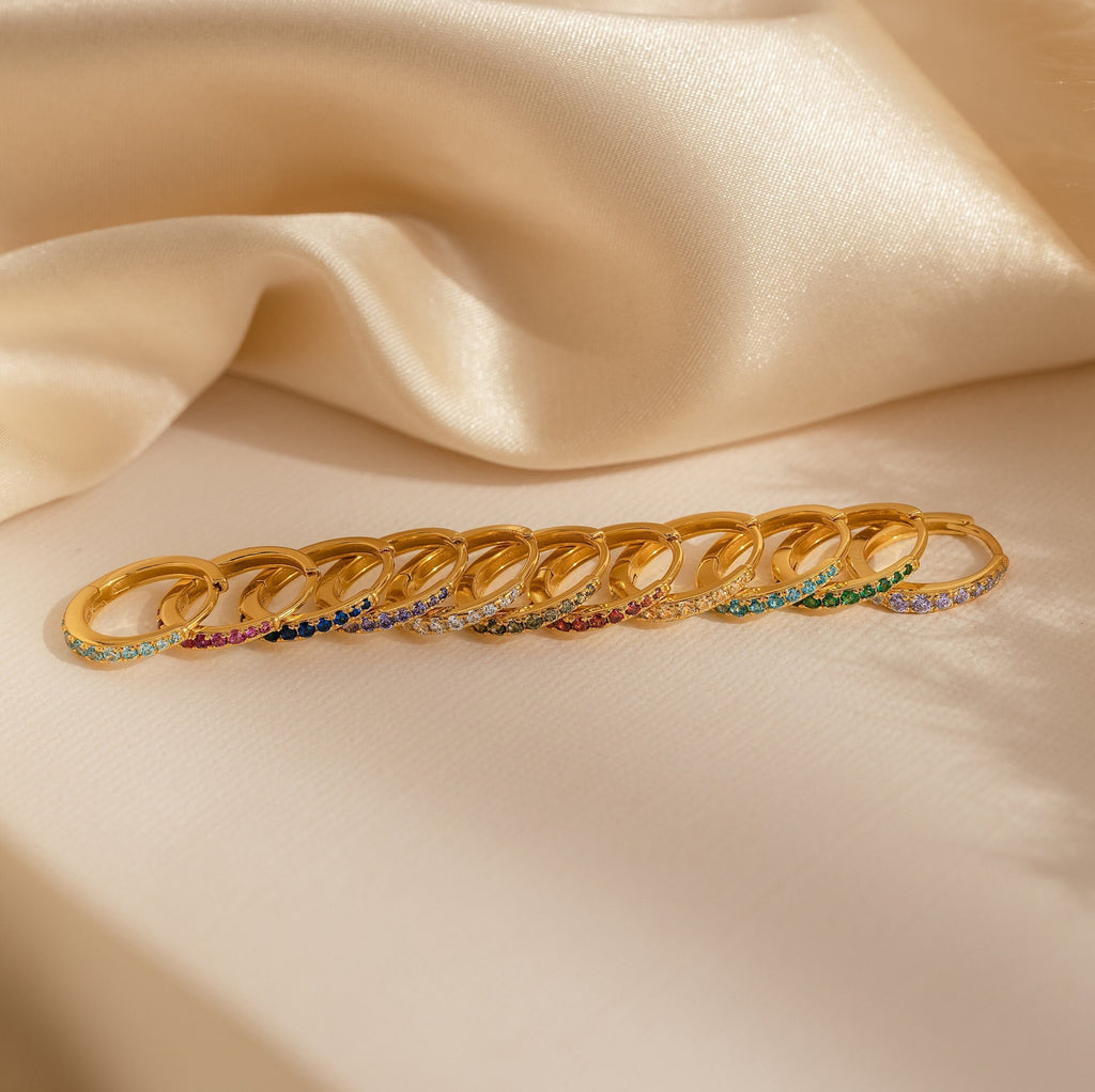 A row of Vera Ruby Birthstone Huggies with vibrant gemstones displayed on cream-colored satin fabric.