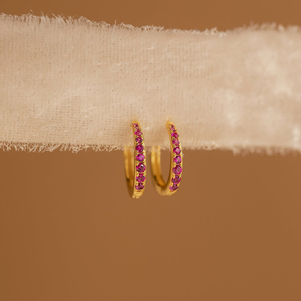 The Vera Ruby Birthstone Huggies feature gold hoops with small ruby stones, shown on textured white fabric against a brown background—these vibrant earrings add elegance to any outfit.