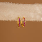 The Vera Ruby Birthstone Huggies feature gold hoops with small ruby stones, shown on textured white fabric against a brown background—these vibrant earrings add elegance to any outfit.