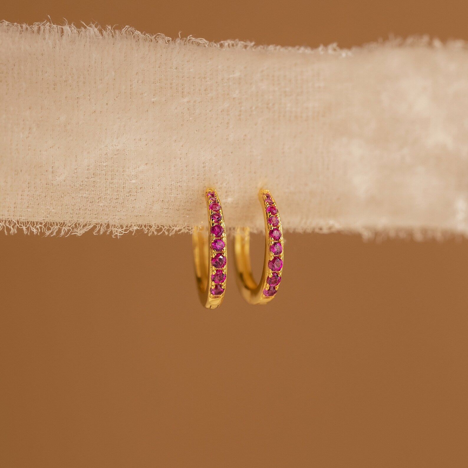 The Vera Ruby Birthstone Huggies feature gold hoops with small ruby stones, shown on textured white fabric against a brown background—these vibrant earrings add elegance to any outfit.