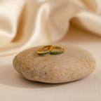 The Vera Emerald Birthstone Huggies, a pair of elegant earrings, are displayed on a rock.