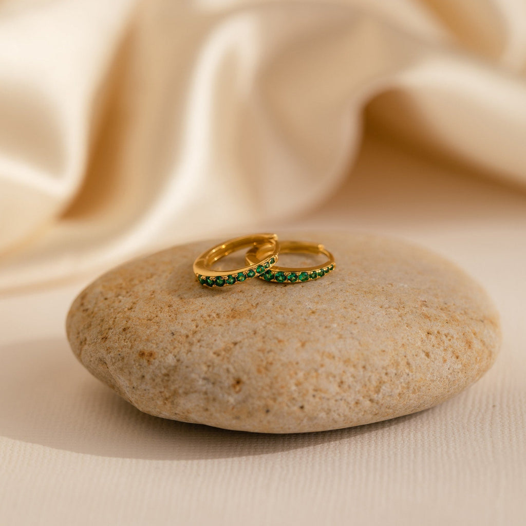 The Vera Pave Birthstone Huggies, a vibrant pair of gemstone earrings, are elegantly displayed on a rock.
