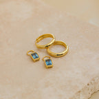 Two Dangling Gemstone Hoops with blue gemstones rest on a beige stone surface.