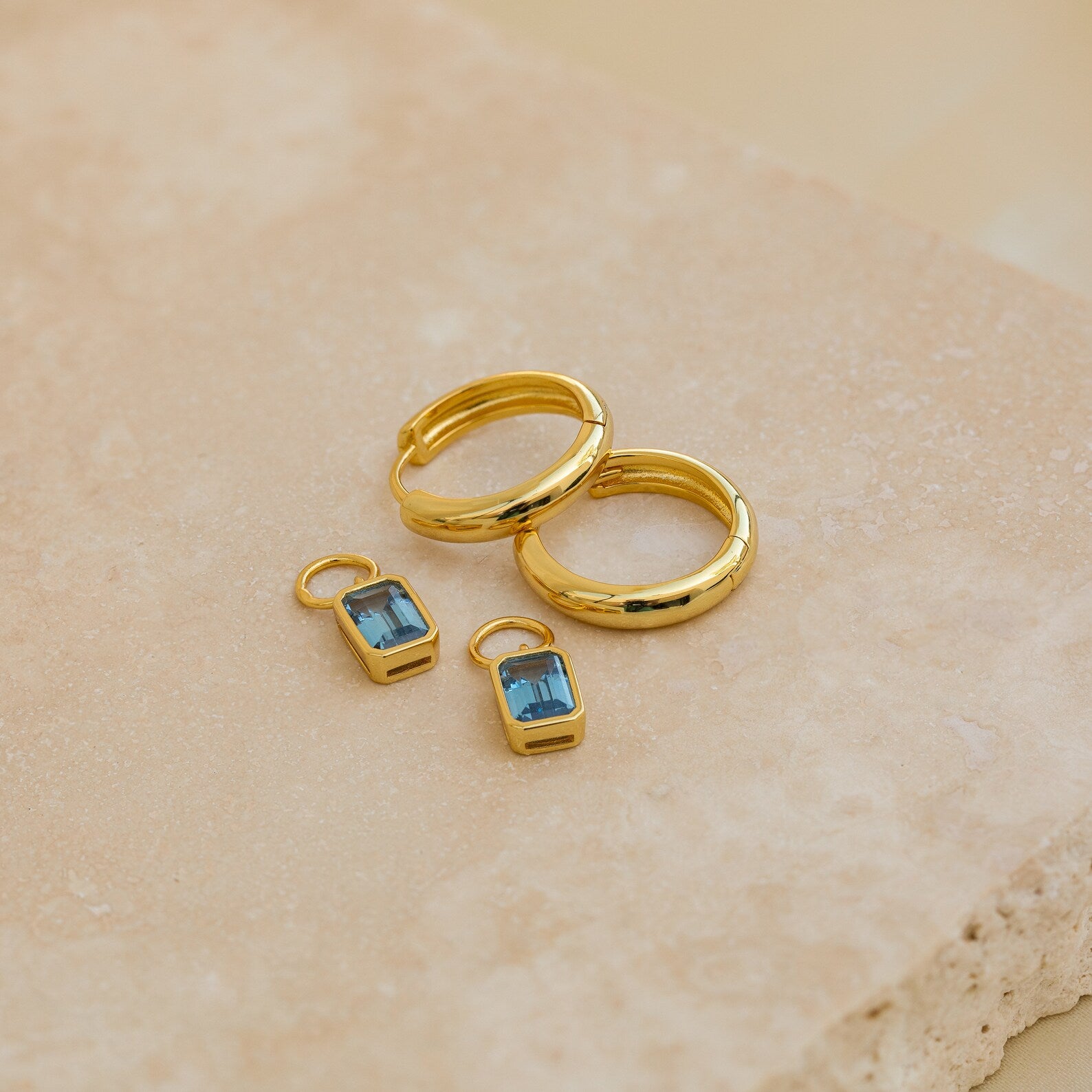 Two Dangling Gemstone Hoops with blue gemstones rest on a beige stone surface.