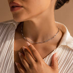 A woman wearing the Diamond Paperclip Necklace with a white textured blouse gently touches her collarbone, creating a chic statement jewelry look.