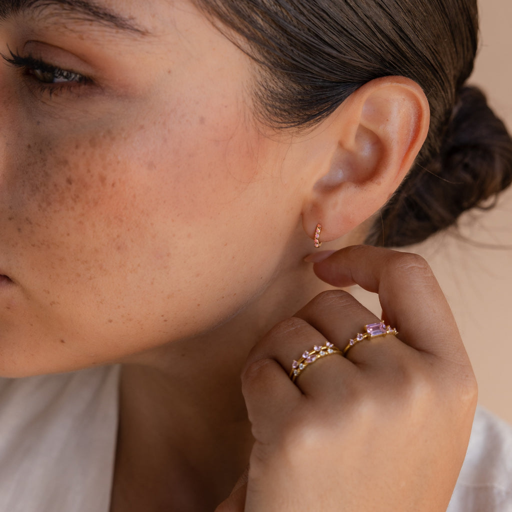 A woman with brown hair in a bun wears Pink Opal Huggies and delicate gold rings with pink stones, her hand near her face—an elegant display of feminine jewelry.