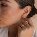 A woman with brown hair in a bun wears Pink Opal Huggies and delicate gold rings with pink stones, her hand near her face—an elegant display of feminine jewelry.