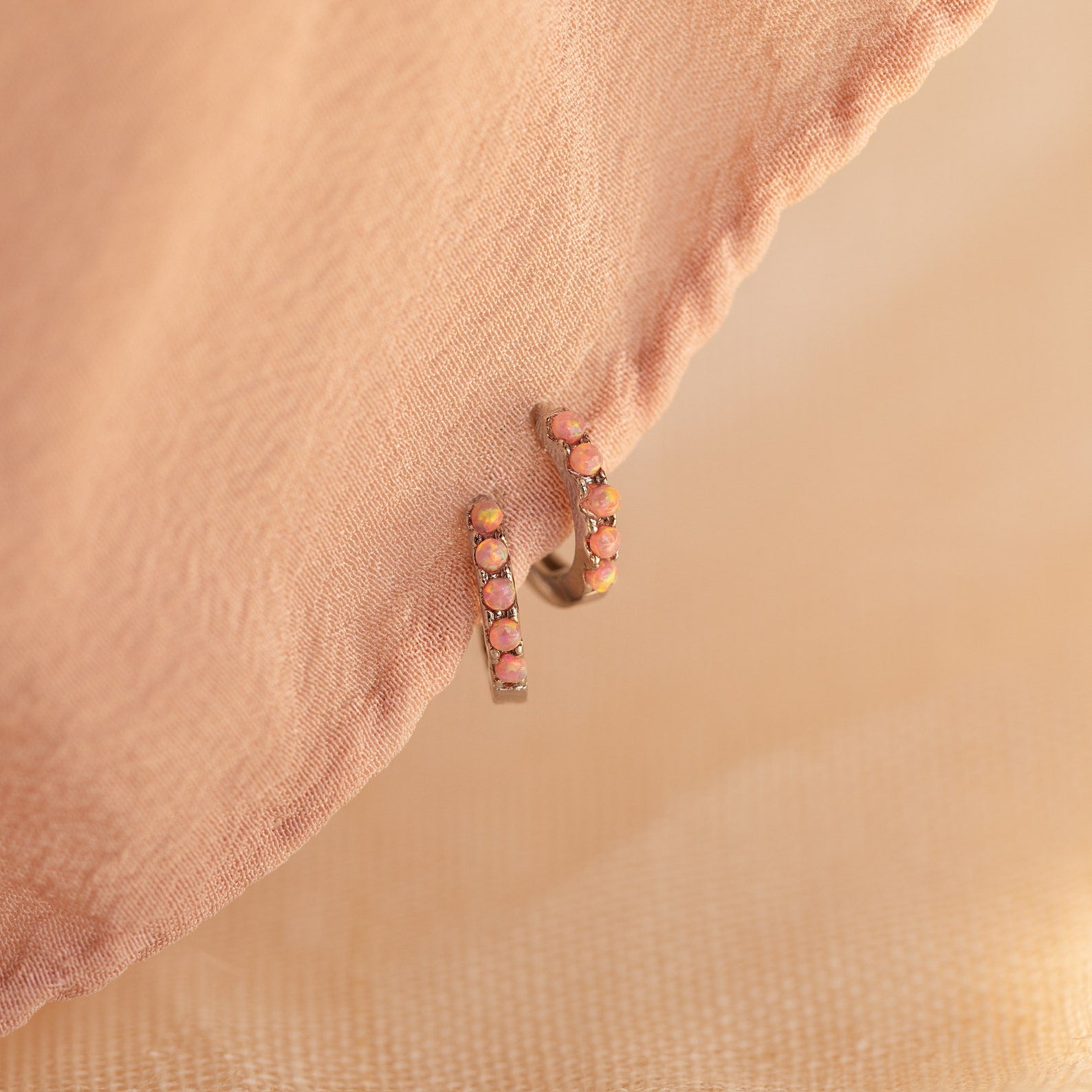 Pink Opal Huggies are gold dainty hoop earrings with small pink opal gems, displayed on soft peach fabric—perfect for adding a feminine touch to any look.