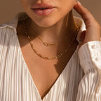 A woman wearing a white pleated blouse and layered gold chain necklaces, including the Diamond Paperclip Necklace, touches her neck to showcase her statement jewelry.
