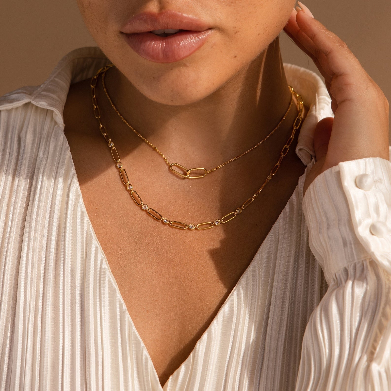 A woman wearing a white pleated blouse and layered gold chain necklaces, including the Diamond Paperclip Necklace, touches her neck to showcase her statement jewelry.