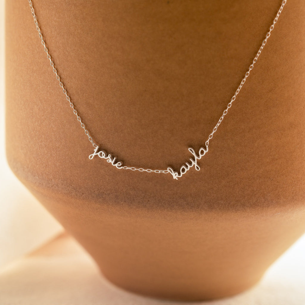 The Dainty Tiny Multiple Name Necklace features josie and kayla in cursive, draped over a tan vase—an ideal personalized accessory.