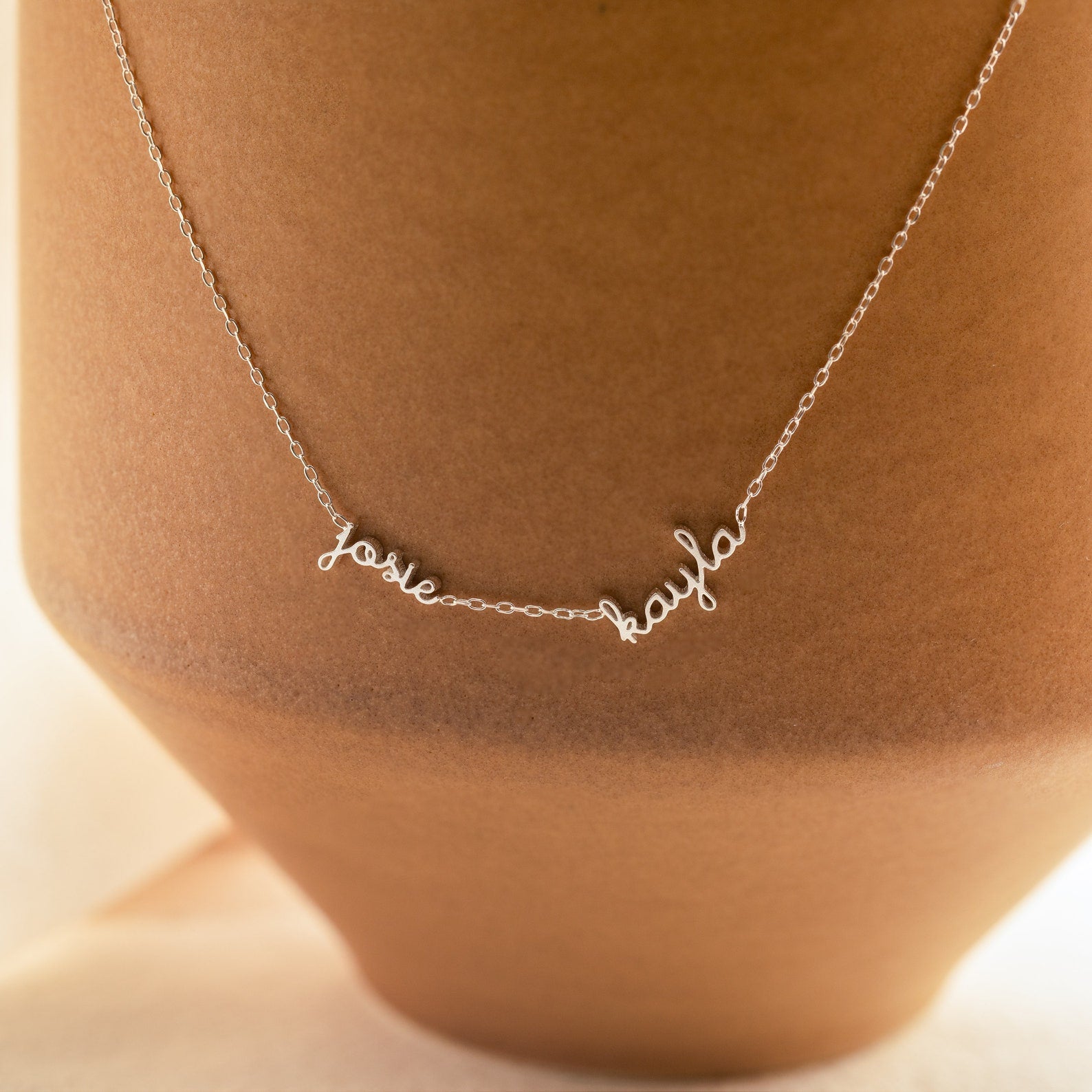 The Dainty Tiny Multiple Name Necklace features josie and kayla in cursive, draped over a tan vase—an ideal personalized accessory.