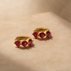 Triple Ruby Birthstone Huggies featuring three vibrant pink rubies displayed on a neutral beige surface—a stunning piece of personalized jewelry.