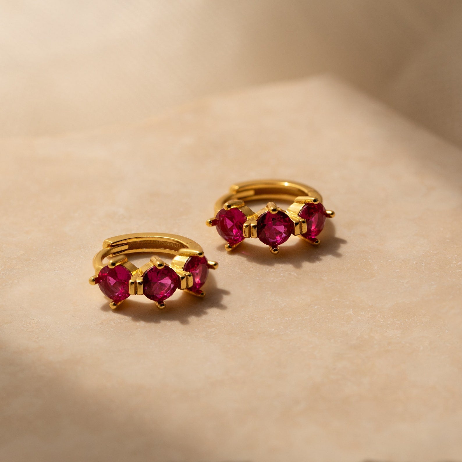 Triple Ruby Birthstone Huggies featuring three vibrant pink rubies displayed on a neutral beige surface—a stunning piece of personalized jewelry.