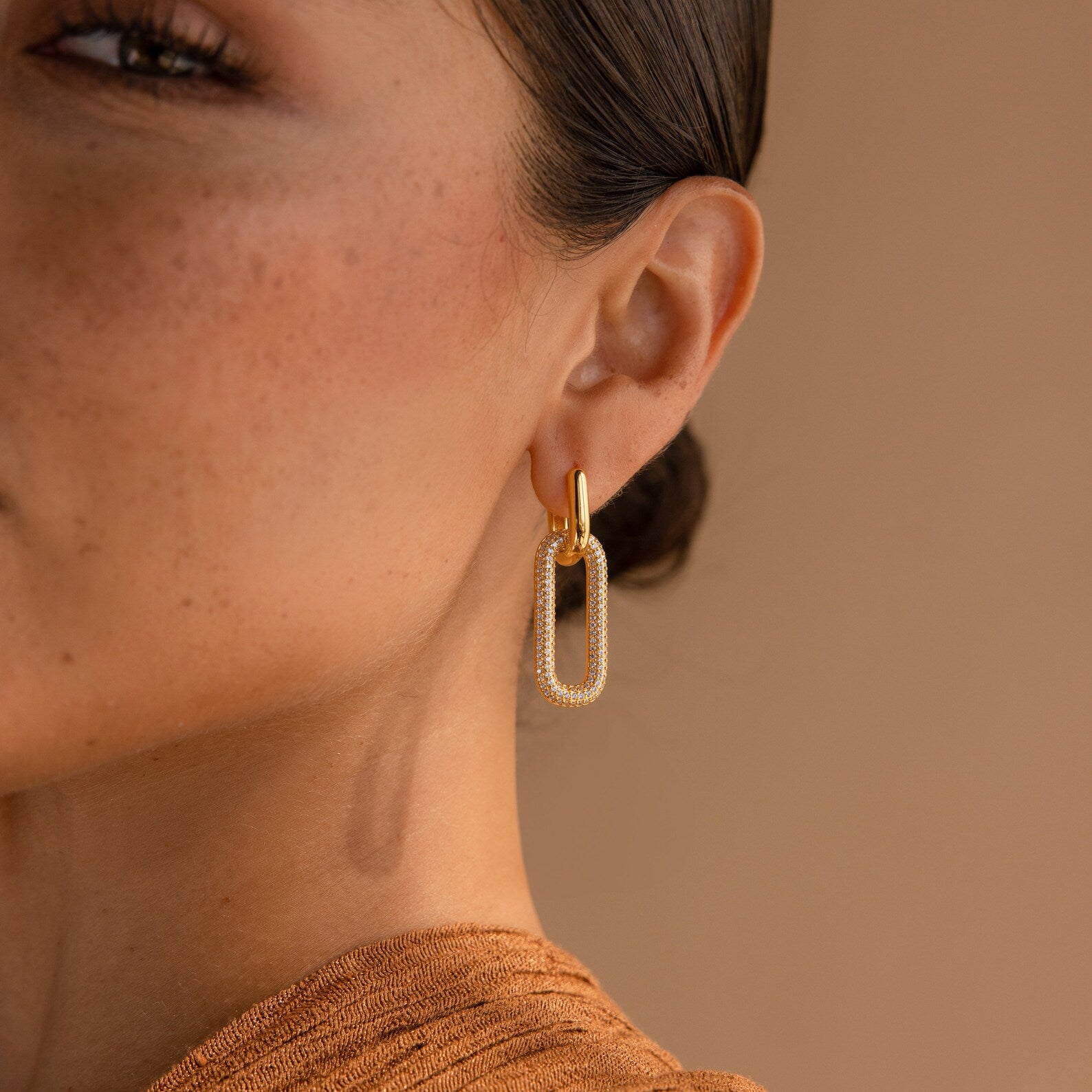 Close-up of a woman's ear wearing Pave Link Hoops—elegant gold earrings with diamond accents—set against a beige background.