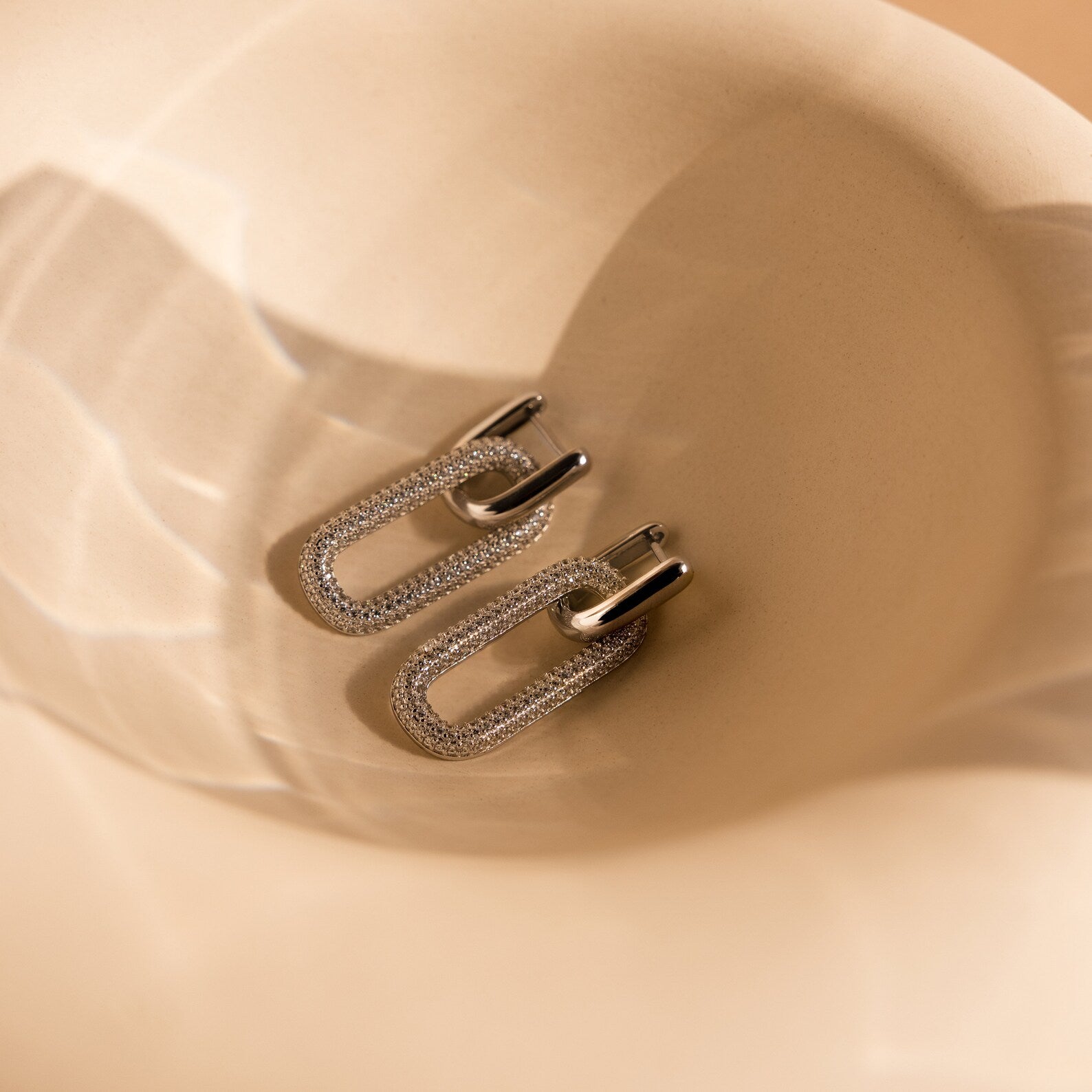 Pave Link Hoops rest on a soft beige, curved surface, catching the light with sparkling reflections from their silver and rhinestone design.