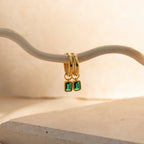 Dangling Gemstone Hoops with a gold finish and removable emerald-cut gemstones elegantly hang on a curved white branch against a beige background, adding sophistication to any outfit.