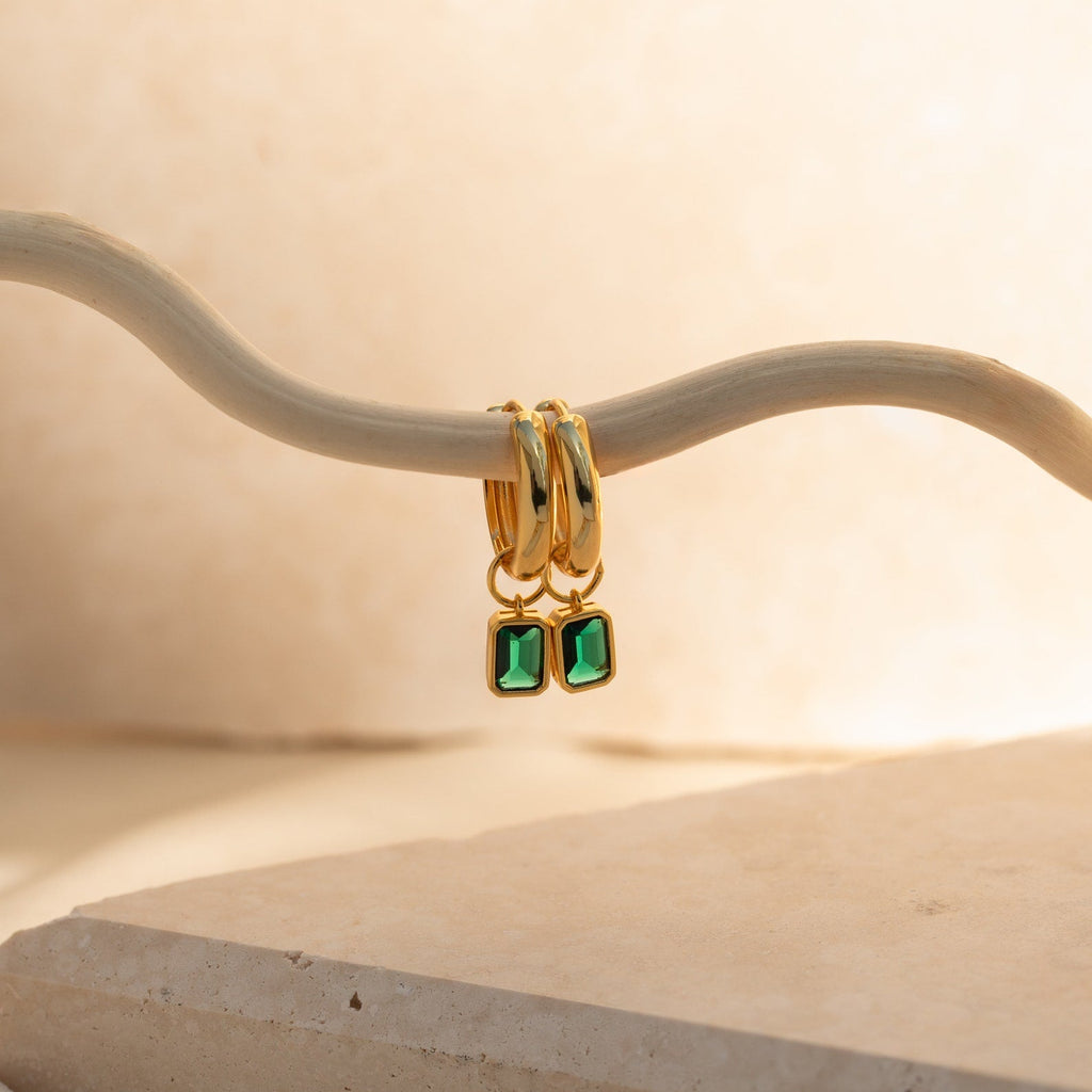 Dangling Emerald Birthstone Hoops with emerald-cut green gemstones hang from a curved beige branch against a soft neutral background; these earrings feature removable stones for versatile styling.