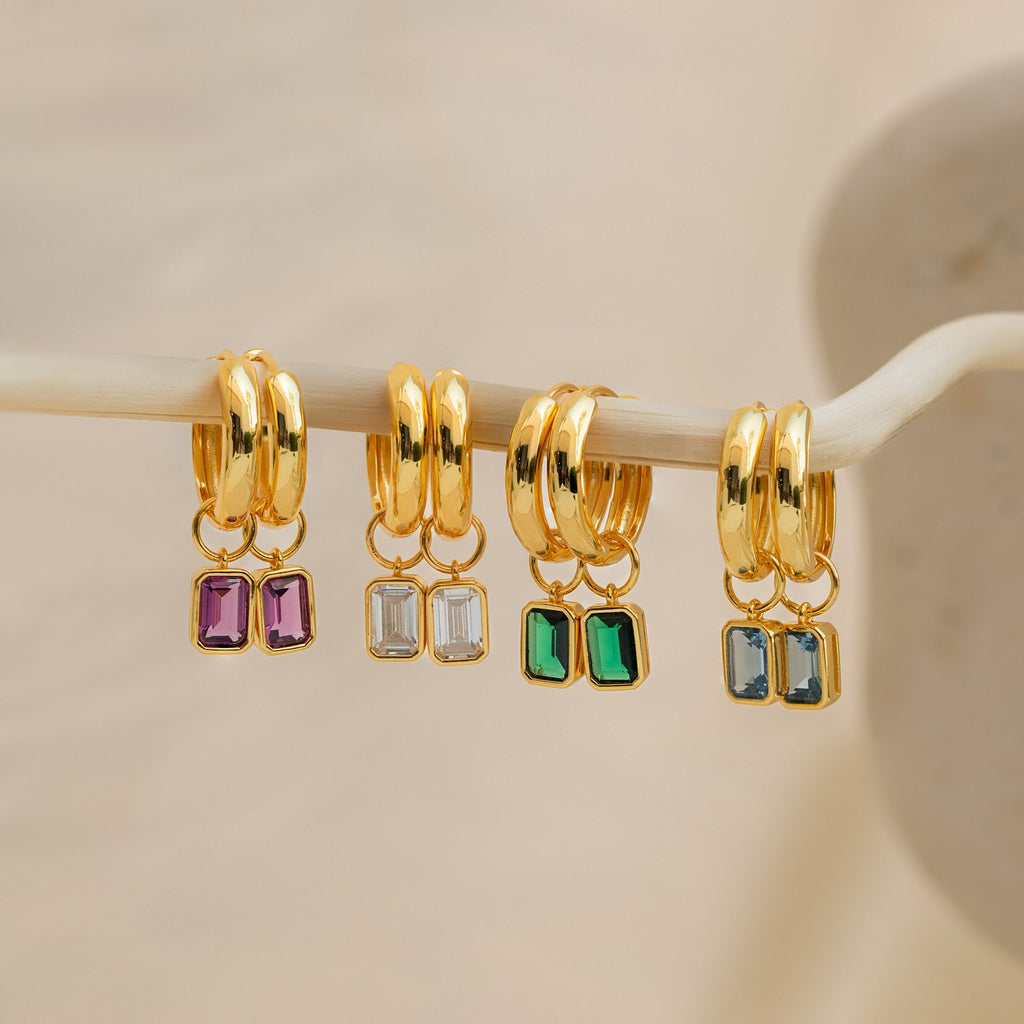 Four pairs of Dangling Gemstone Hoops with gold finish and removable emerald-cut charms in purple, clear, green, and blue add vibrant color to any look. The chic earrings hang from a branch.