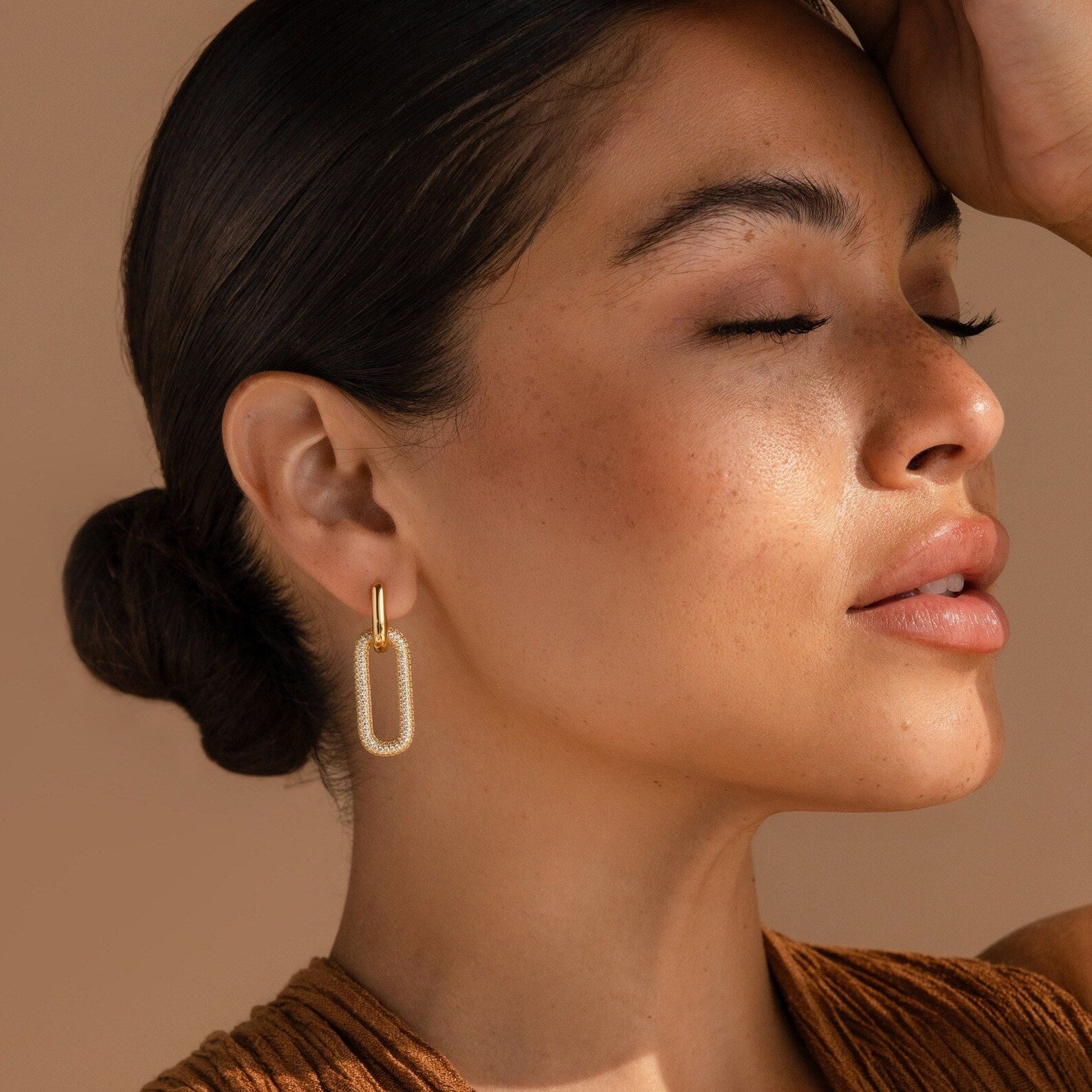 Woman with closed eyes wears Pave Link Hoops, her hair in a low bun and arm raised to her forehead.