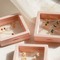 The Birthstone Mystery Box features a gold necklace and earrings with red and green gemstones, elegantly displayed in pink square boxes on a light surface, offering a personalized surprise.