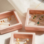 The Birthstone Mystery Box features a gold necklace and earrings with red and green gemstones, elegantly displayed in pink square boxes on a light surface, offering a personalized surprise.