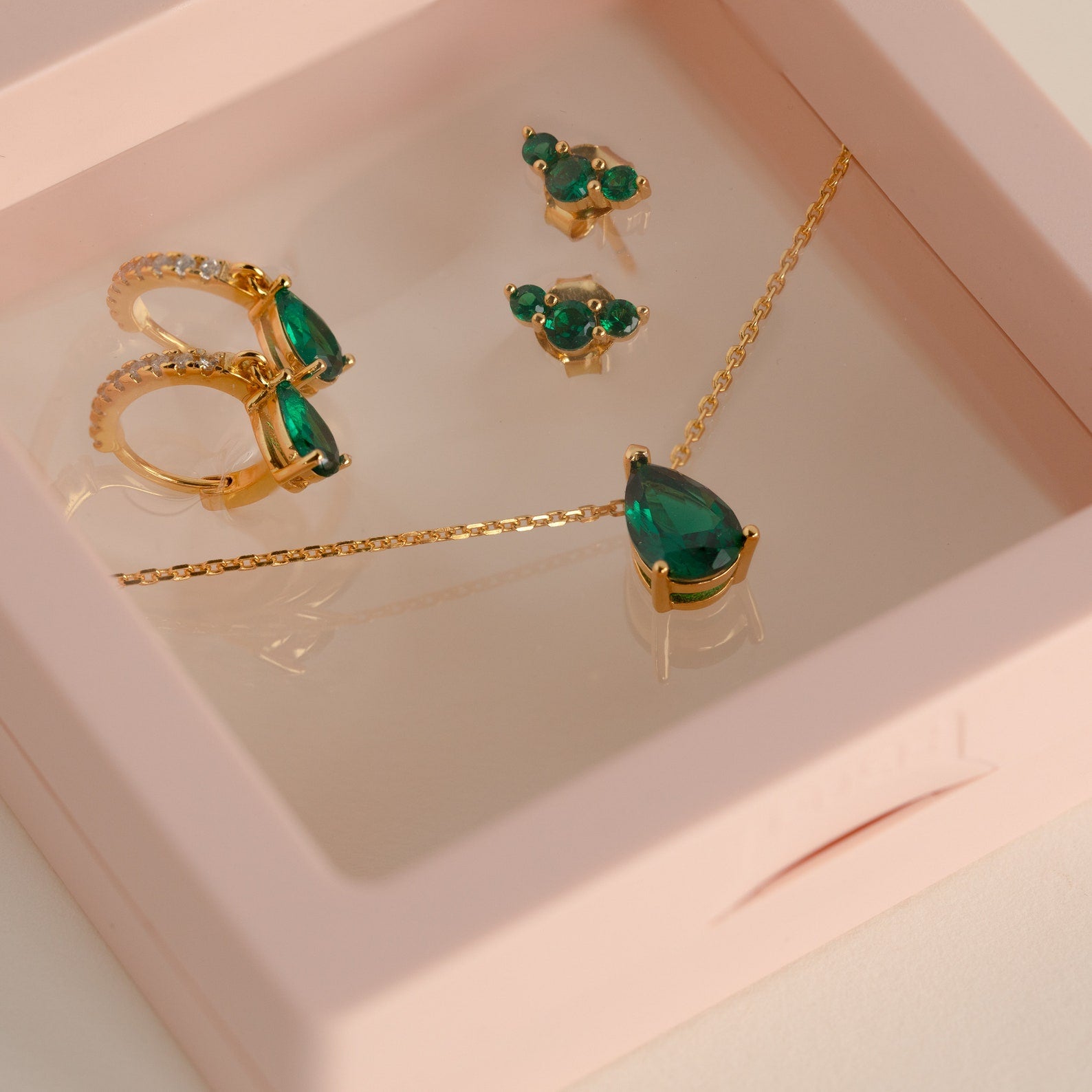 The Birthstone Mystery Box features gold jewelry with green gemstones in a pink box, including a necklace, earrings, rings, and a teardrop pendant—ideal for gifting or a personalized surprise.