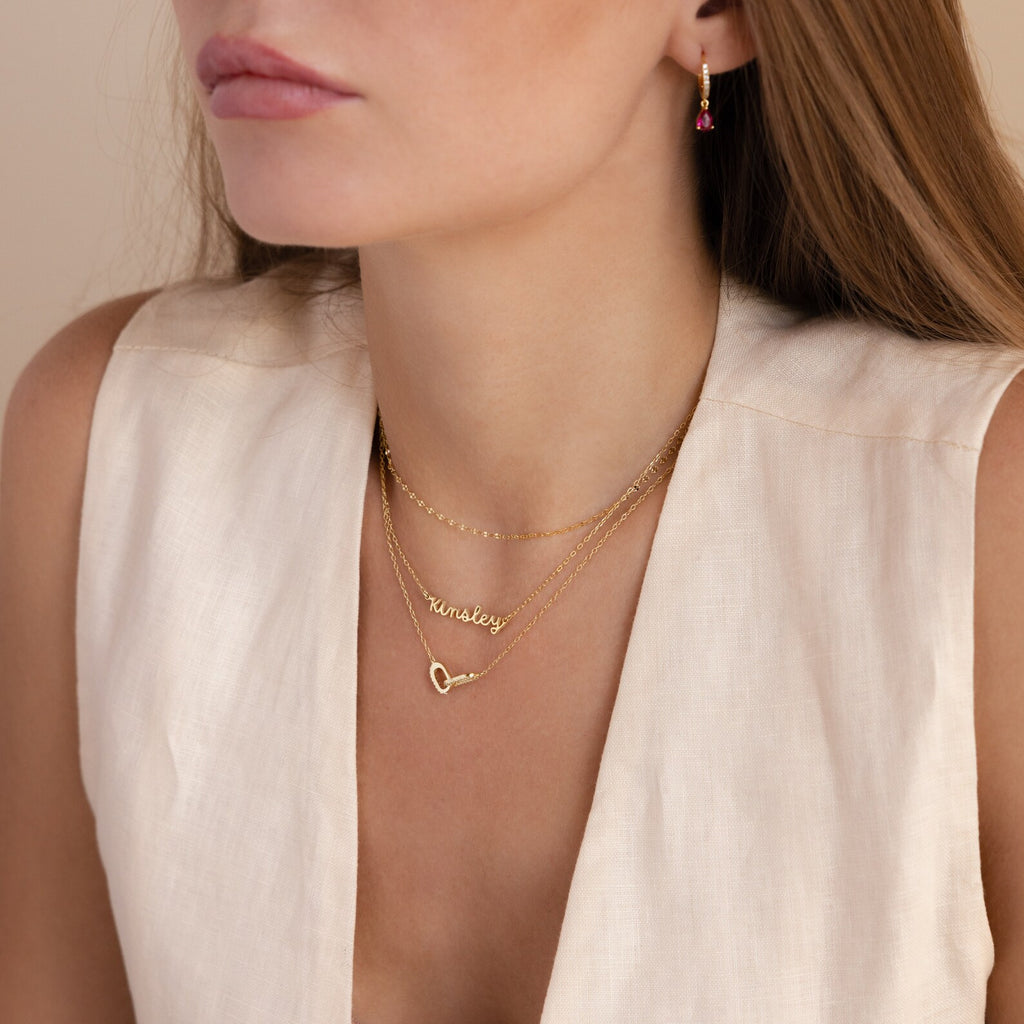 A woman wears layered gold necklaces, including the Tiny Dream Name Necklace featuring a cursive font and heart pendant, paired with a sleeveless beige top.