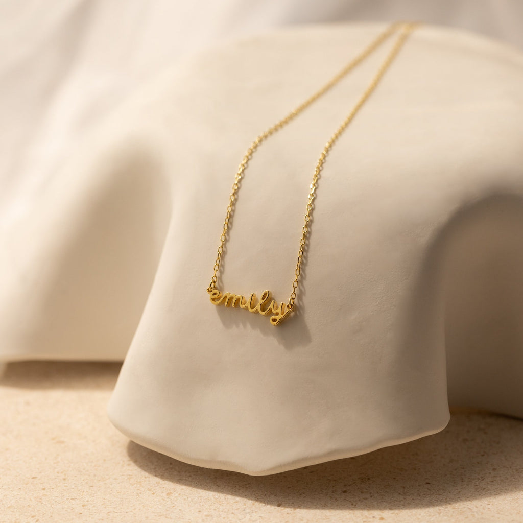 The Tiny Dream Name Necklace showcases "Emily" in graceful cursive on soft, light fabric—this personalized piece adds a delicate, stylish accent to any outfit.