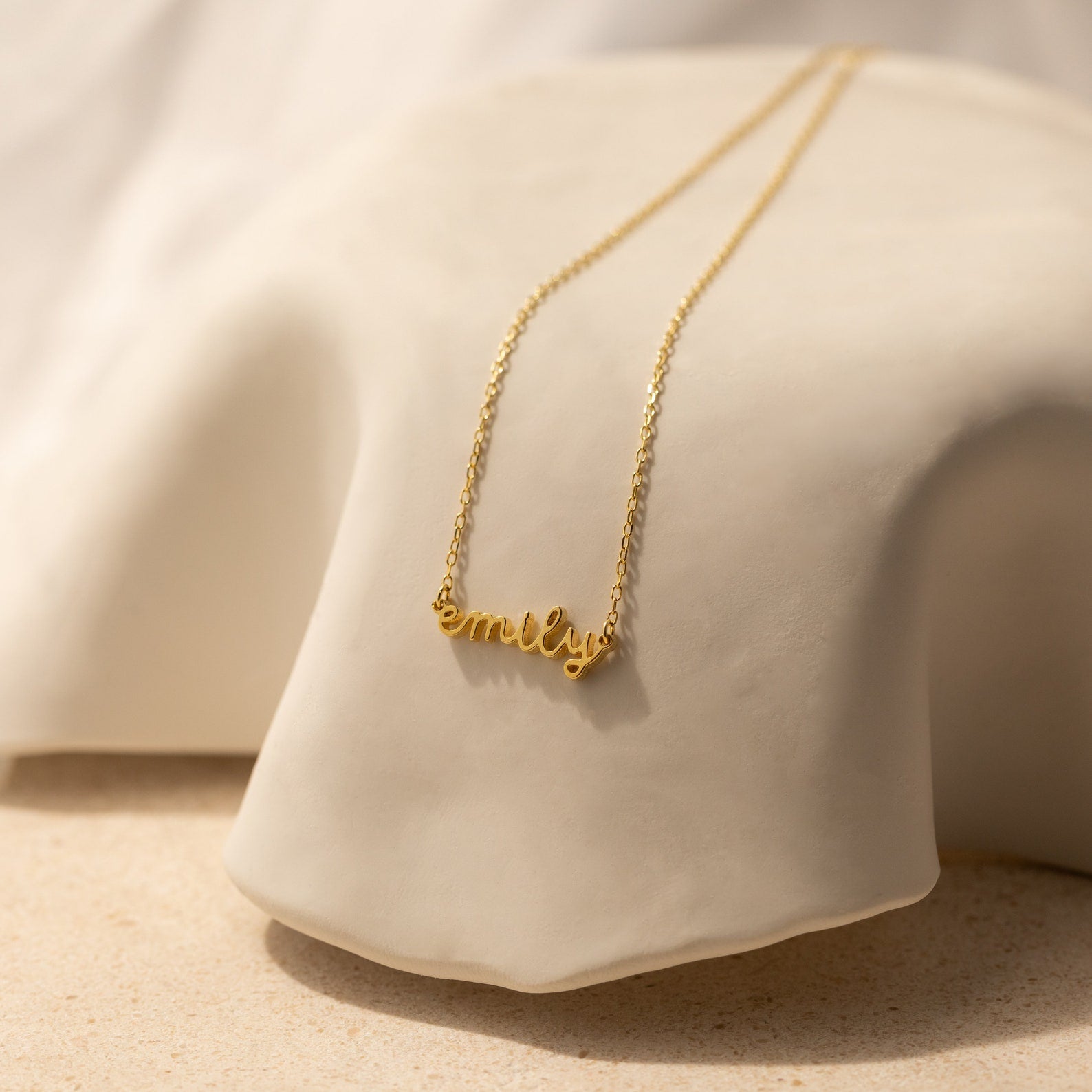 The Tiny Dream Name Necklace showcases "Emily" in graceful cursive on soft, light fabric—this personalized piece adds a delicate, stylish accent to any outfit.
