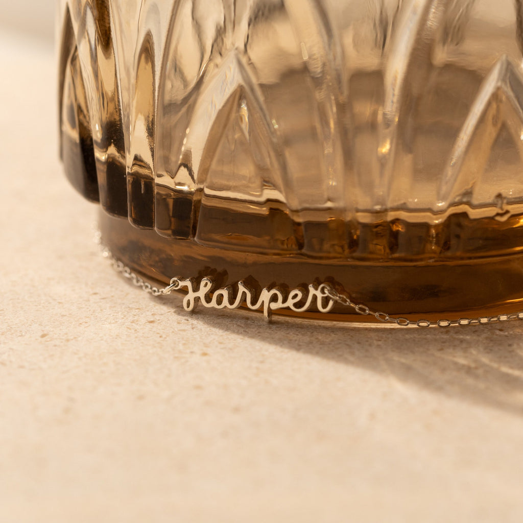 The Tiny Dream Name Necklace, featuring "Harper" in delicate cursive, rests at the base of a textured glass container.