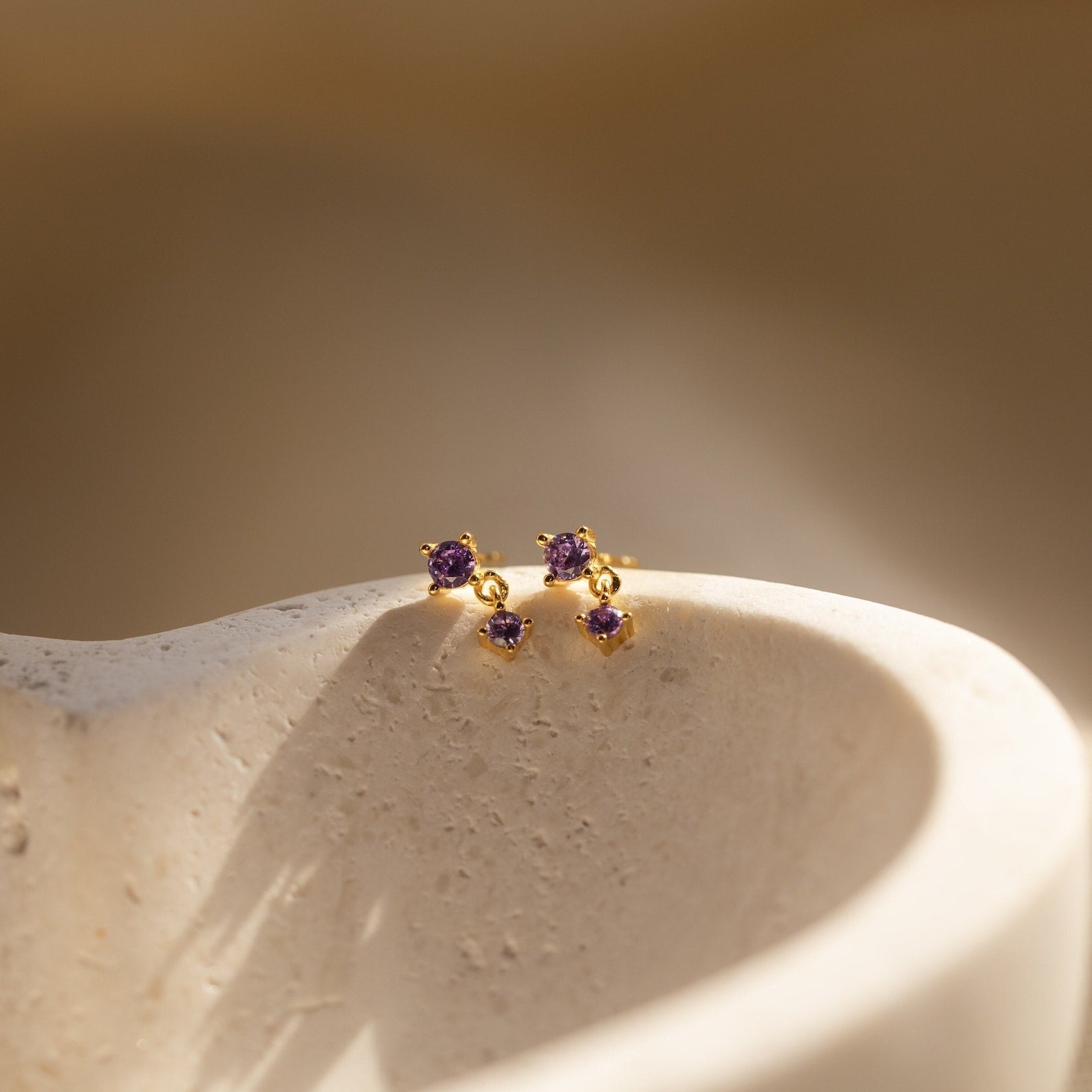 Opal Birthstone Drop Studs with delicate purple gems displayed on a smooth, curved beige stone in soft light—perfect dainty earrings for elegant birth month jewelry.