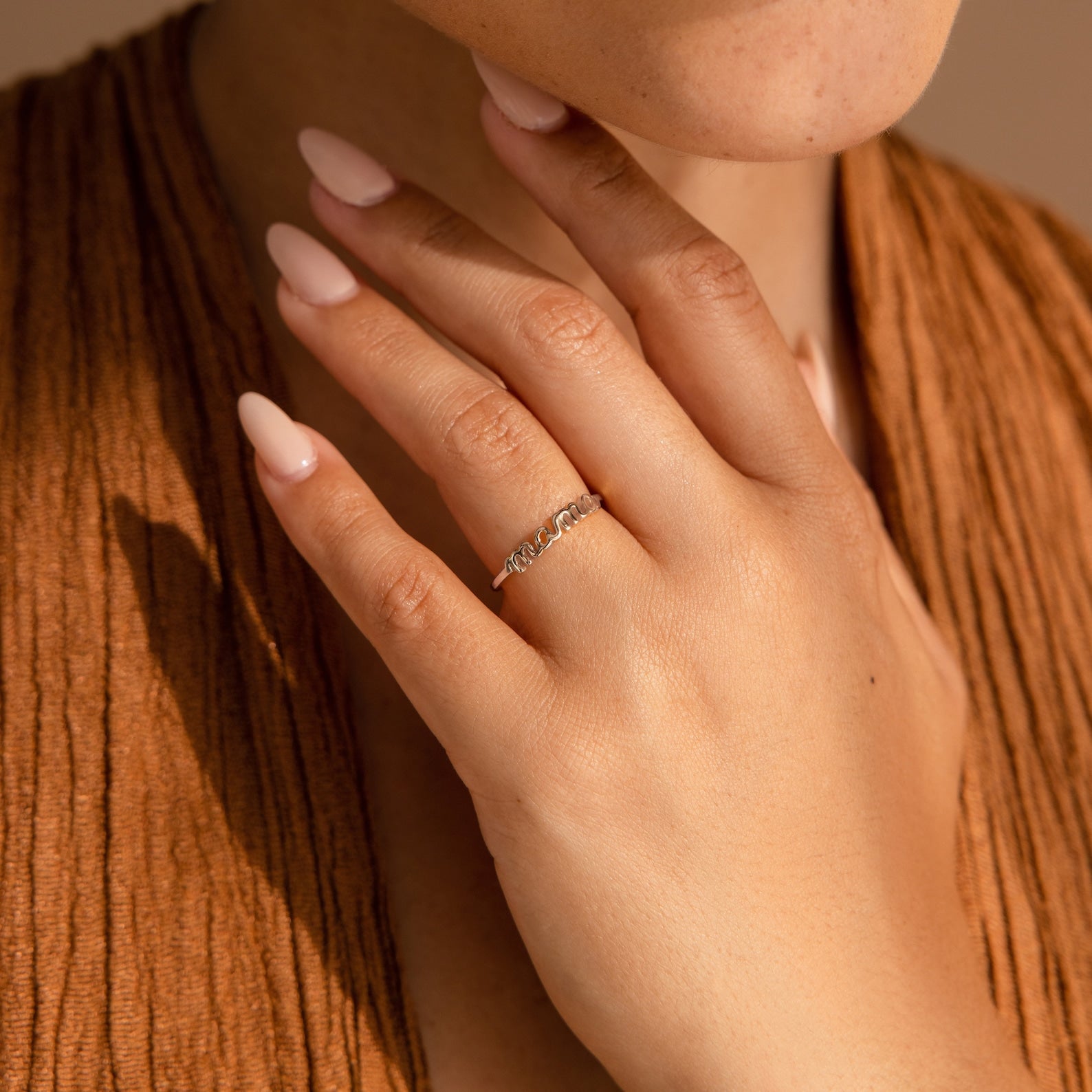 A hand with nude nails wearing the Dainty Mama Ring rests on the chest in warm lighting—a perfect gift for mothers.