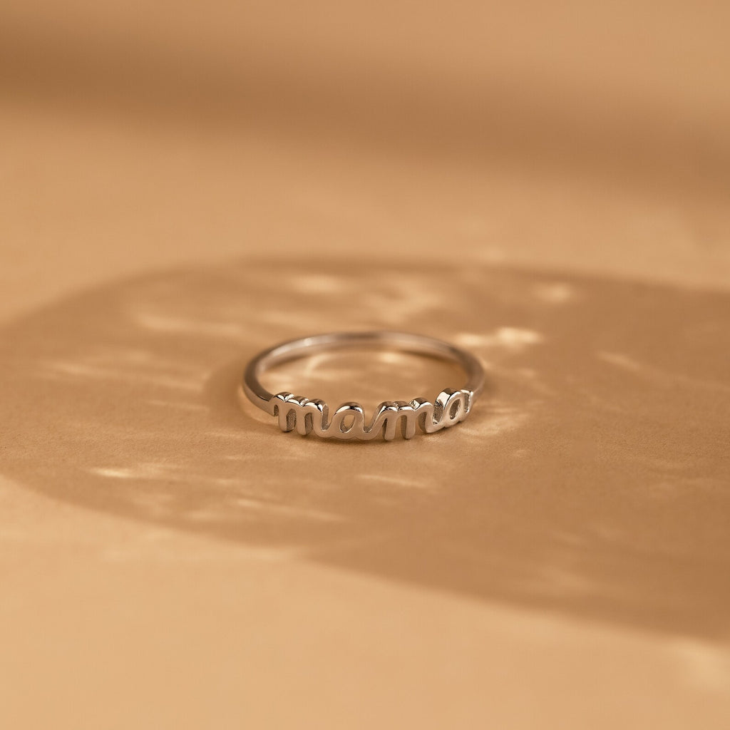 The Dainty Mama Ring, engraved with "mama," rests on a light brown surface with soft shadows—a minimalist and meaningful gift for any mother.
