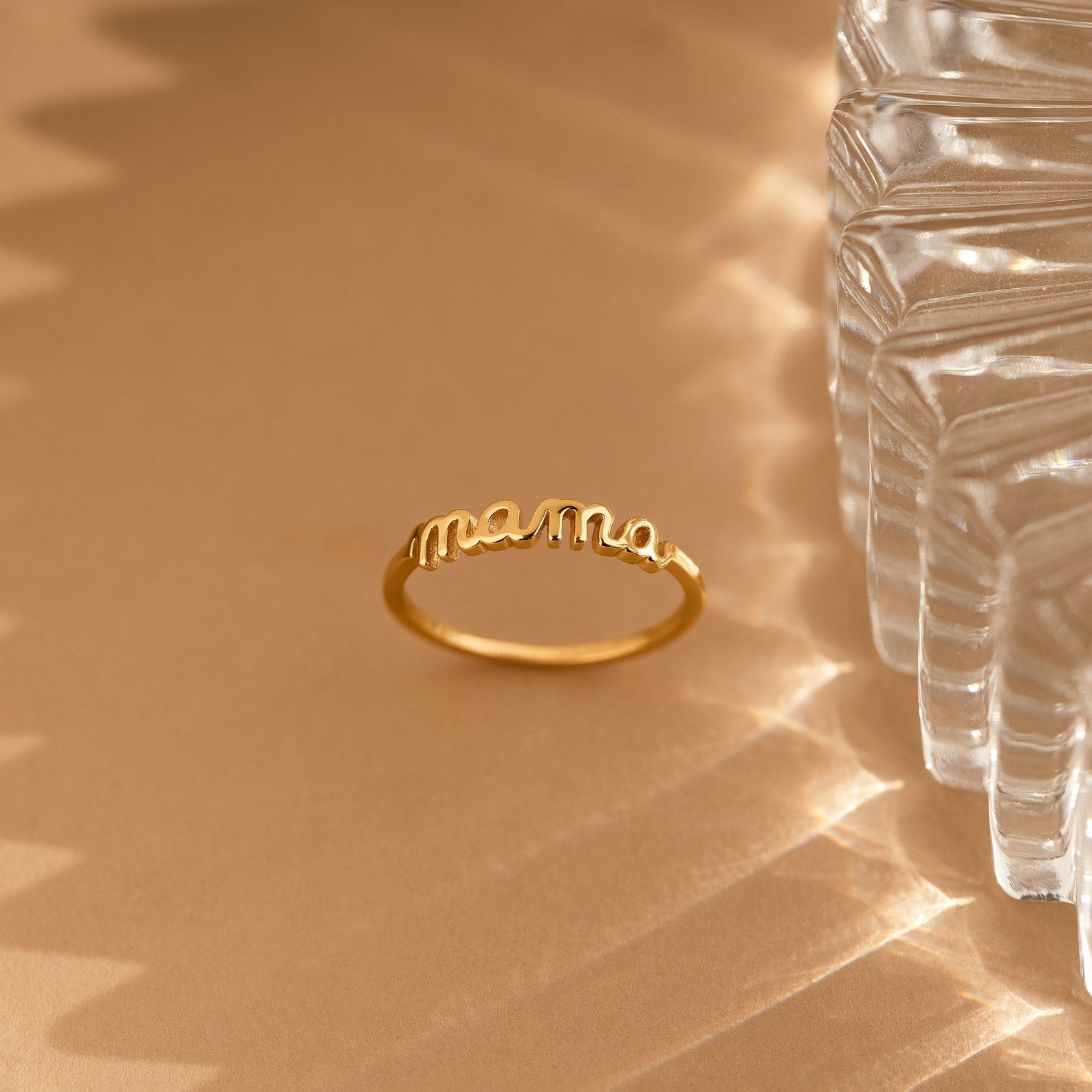The Dainty Mama Ring showcases mama in elegant cursive, displayed on a beige surface next to a textured glass—making it a perfect, thoughtful gift for mom.