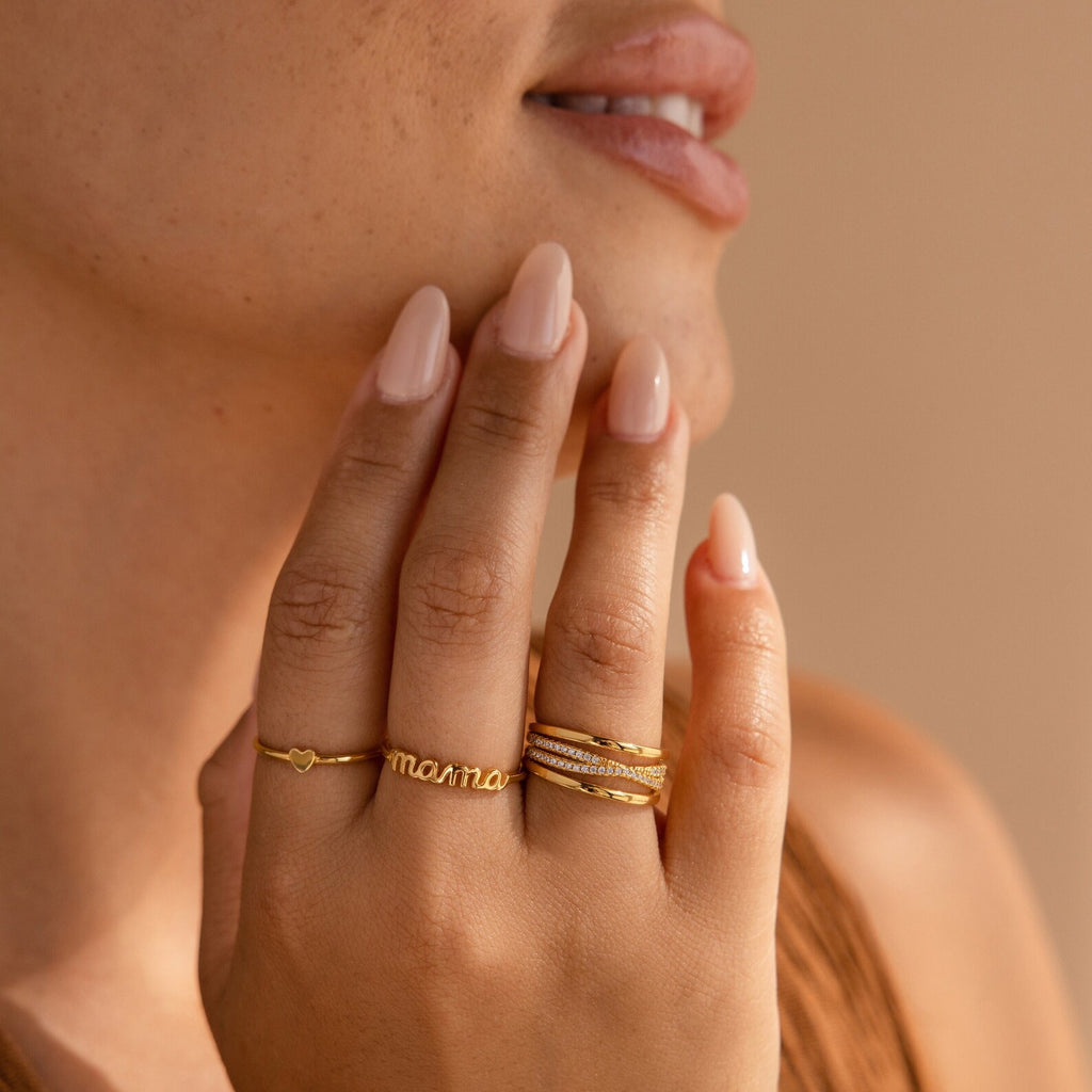 A person with nude manicured nails and dainty jewelry wears multiple gold rings, including the Dainty Mama Ring, holding their hand to their face—a lovely gift idea for mom.