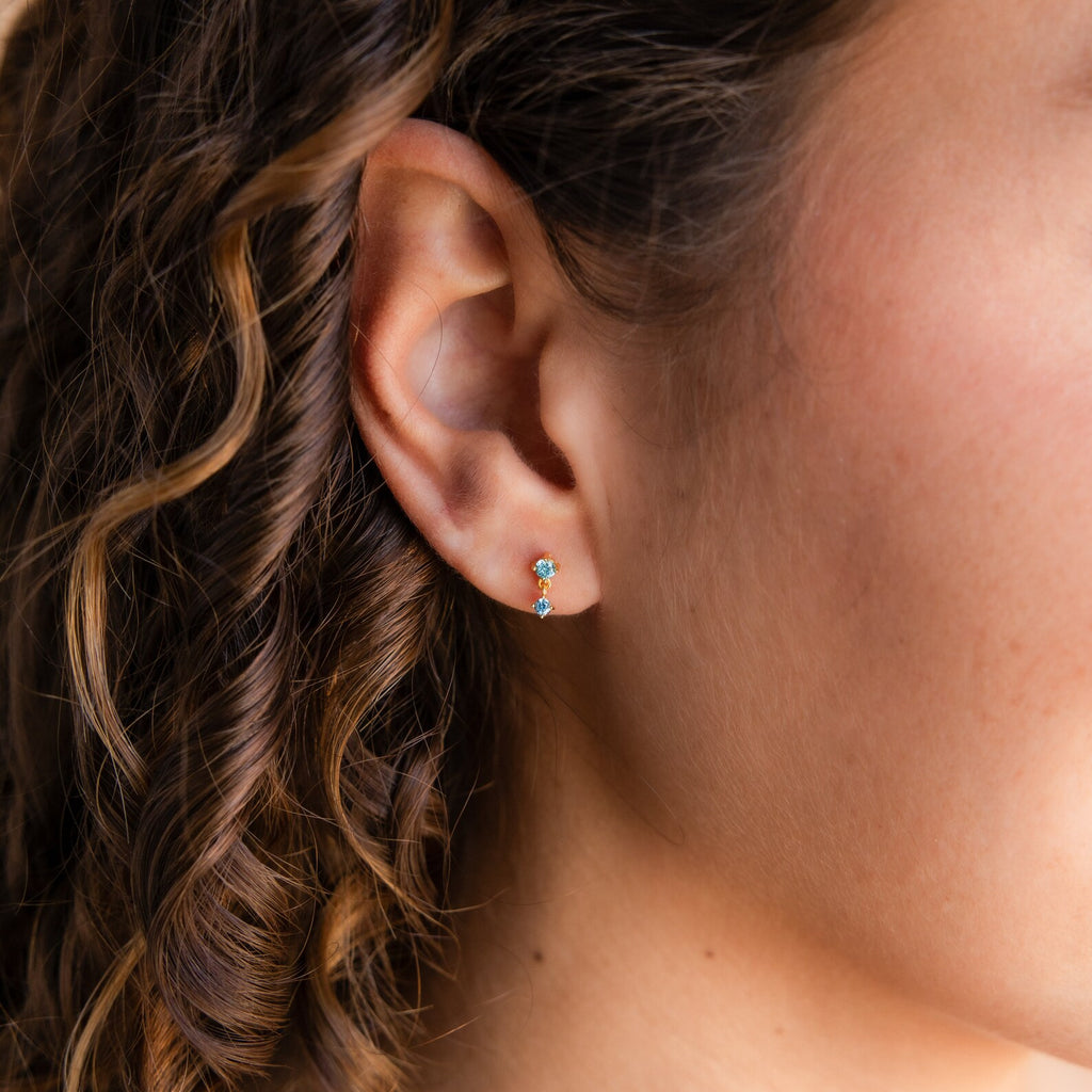 Close-up of a woman's ear wearing Birthstone Drop Studs with blue gemstones; her curly brown hair is visible.