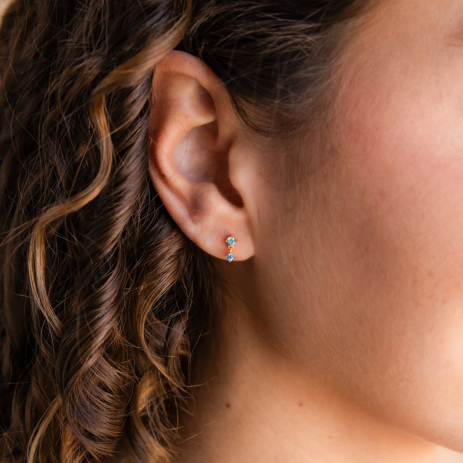 Close-up of a woman's ear with curly brown hair, wearing Opal Birthstone Drop Studs—dainty earrings often chosen as birth month jewelry.