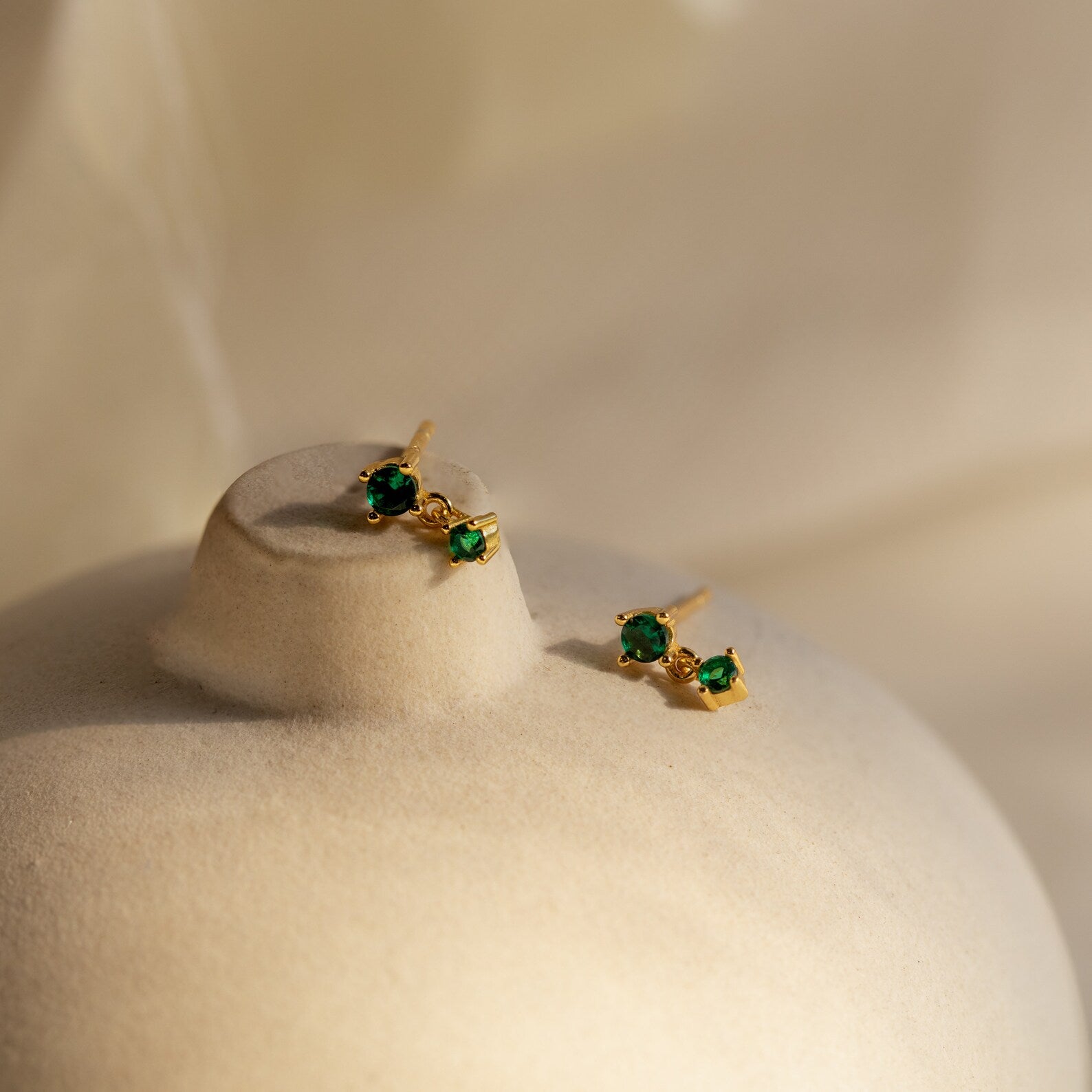 The Birthstone Drop Studs, adorned with small green gemstones, are showcased on a smooth cream-colored surface in soft lighting—perfect dainty earrings to celebrate your birth month.