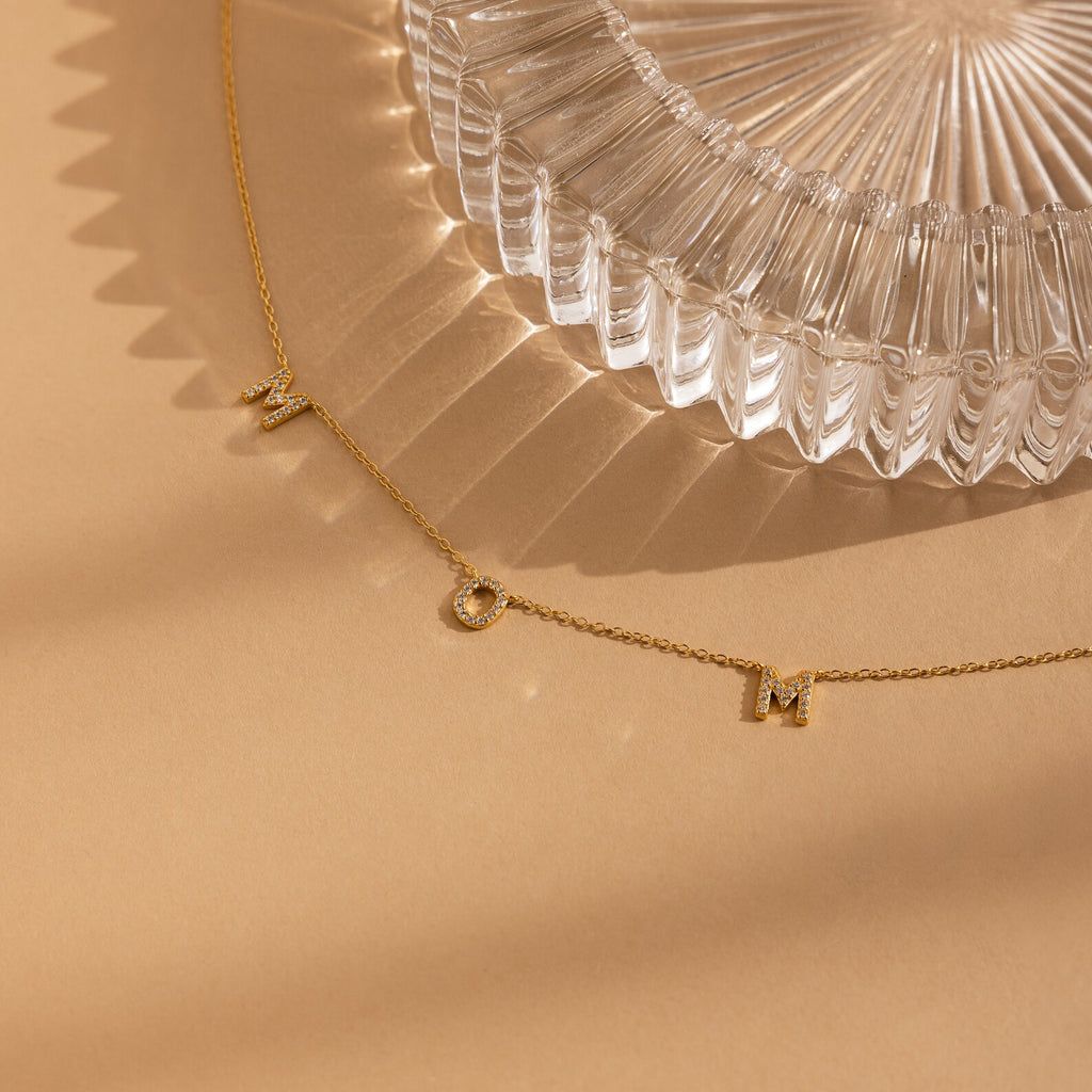 The Pave Mom Necklace, with sparkling letter charms, is shown next to a clear ribbed glass dish on a beige surface—a thoughtful gift that honors mom.