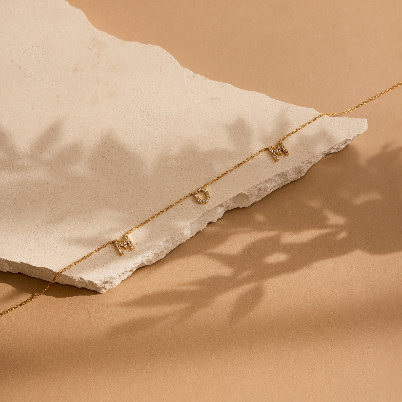 The Pave Mom Necklace, spelling MOM in sparkling pavé, rests on textured beige stone with soft shadows on a tan backdrop—a meaningful gift celebrating a mother's love.