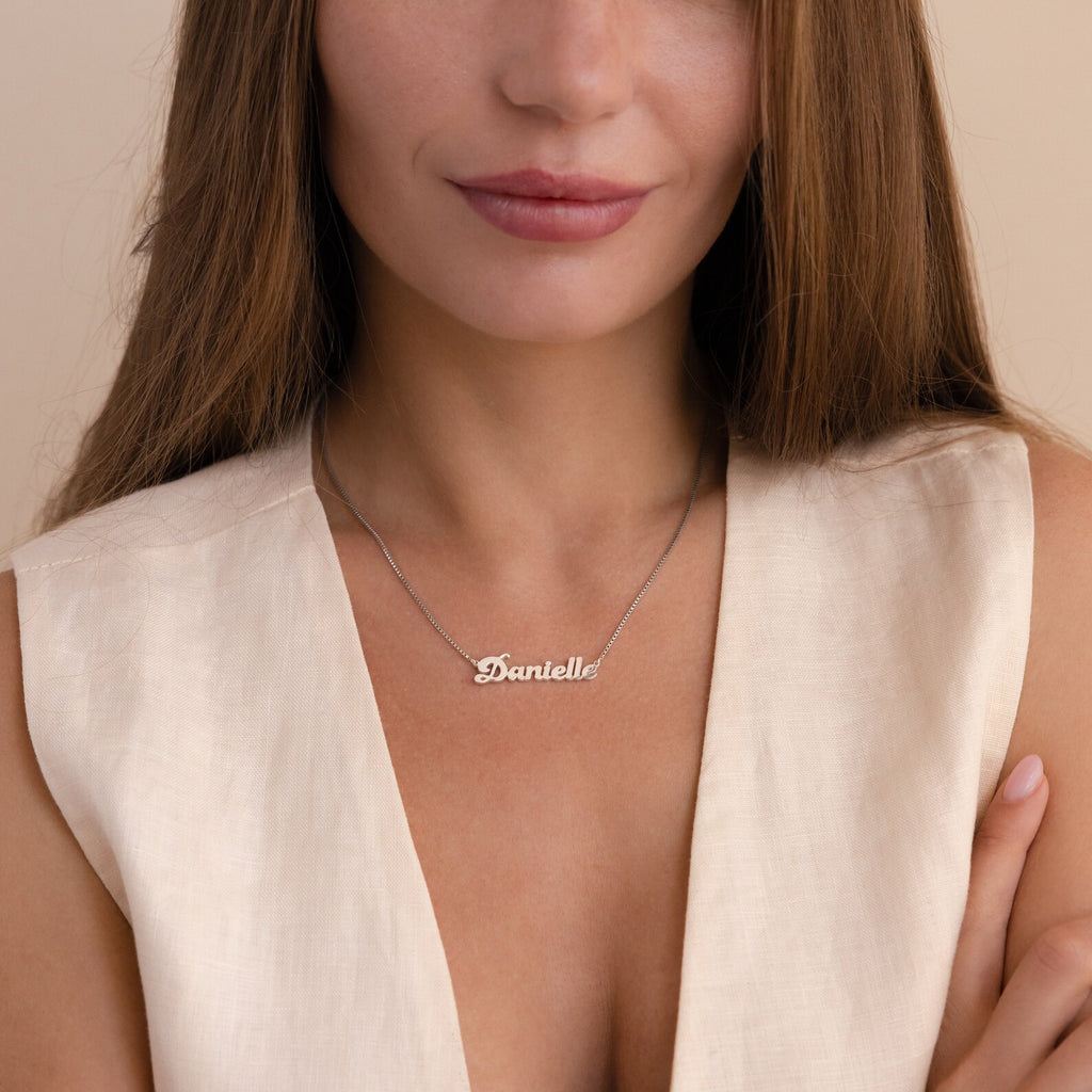 A woman wears a cream sleeveless top and the Groovy Name Necklace in Box Chain, personalized with “Danielle” elegantly written in cursive letters.