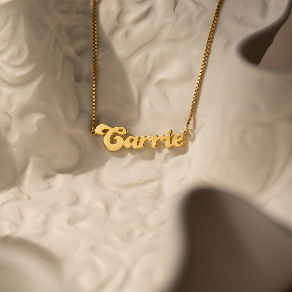 The Groovy Name Necklace in Box Chain, personalized with the name Carrie, rests on a textured white surface and highlights elegant custom jewelry craftsmanship.