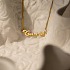 The Groovy Name Necklace in Box Chain, personalized with the name Carrie, rests on a textured white surface and highlights elegant custom jewelry craftsmanship.