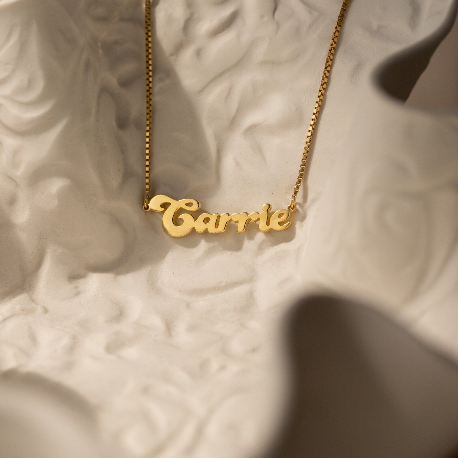 The Groovy Name Necklace in Box Chain, personalized with the name Carrie, rests on a textured white surface and highlights elegant custom jewelry craftsmanship.