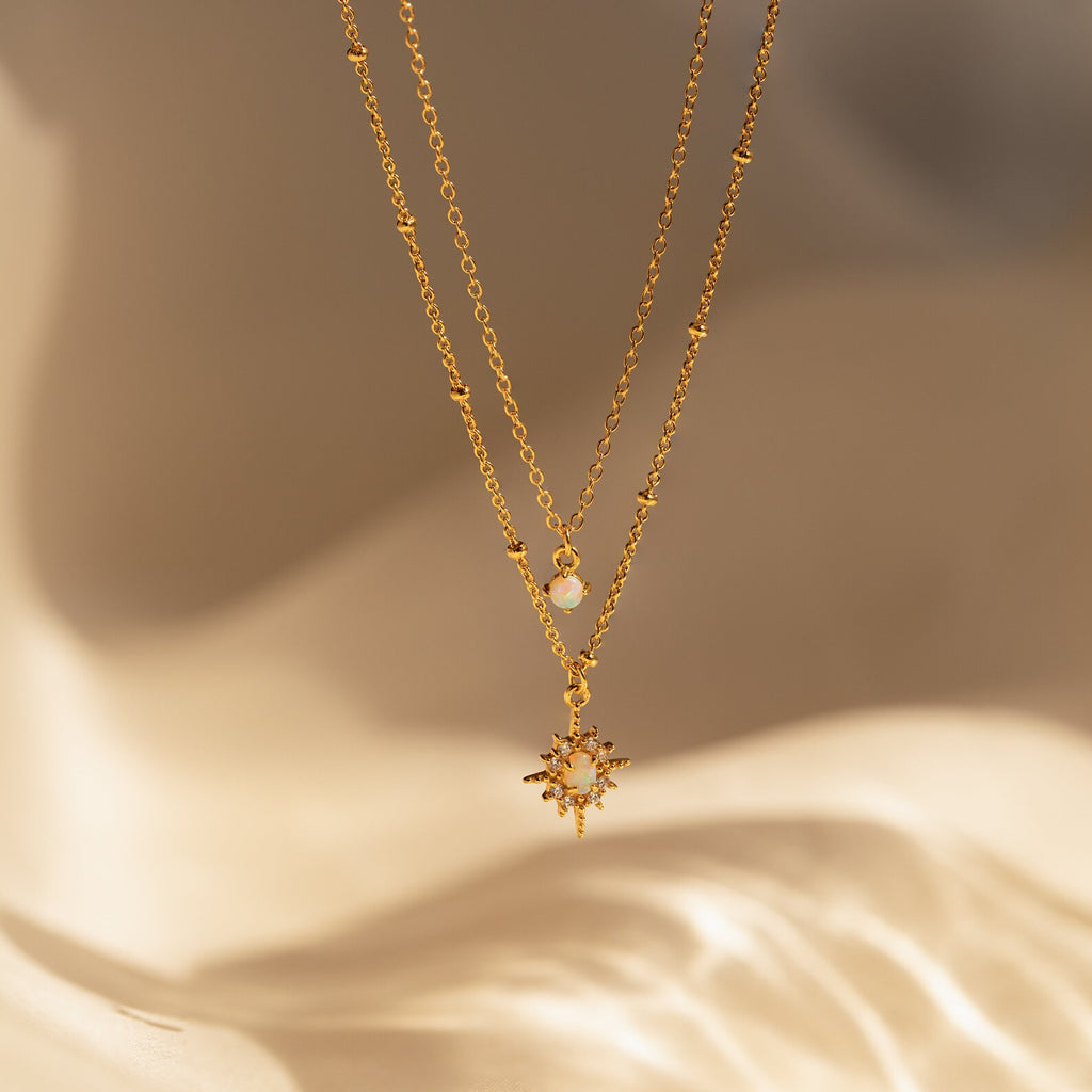 The Opal Star Duo Necklace, featuring a delicate opal and a double chain with starburst design, rests on a soft beige background.